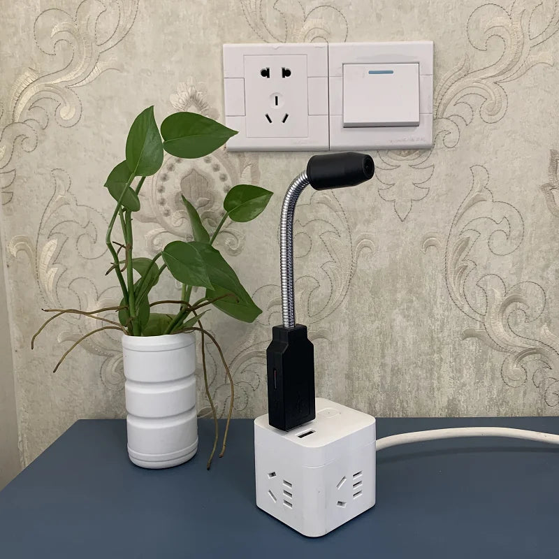 A potted plant with green leaves sits on a blue surface next to a power strip with multiple outlets and USB ports. A flexible USB LED lamp is plugged into the power strip. In the background, there is a wall-mounted power socket and a Mini WiFi Nanny Camera featuring a wide-angle lens.